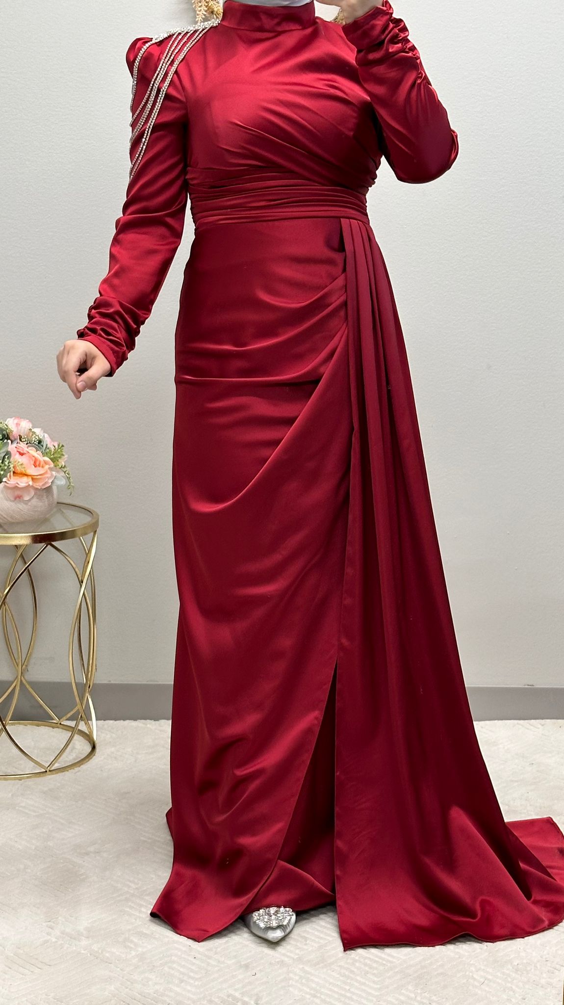 فستان رسمي أحمر مزين بدقة بسلاسل تتدلى على الكتف وطيات متداخلة مصنوعة بدقة. إنه مثال للفخامة، فهو يعرض ثنية جانبية أنيقة، بينما تضفي الأكمام المنتفخة سحرًا ملكيًا.